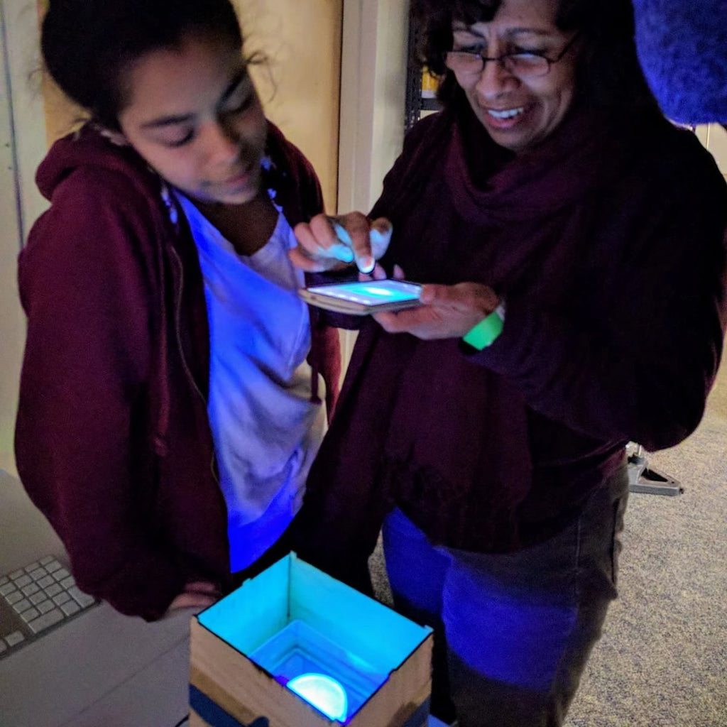 mother and daughter taking a photo of glowing bacteria during a Genetic Engineering DIY biohacking workshop by Amino labs STEM biotechnology kits and school laboratory equipment / home laboratory equipment / biohacking laboratory equipment